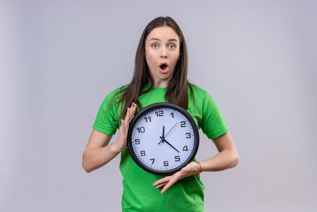 Hermosa joven vestida con camiseta verde sosteniendo el reloj mirando asombrado y sorprendido de pie sobre fondo blanco aislado