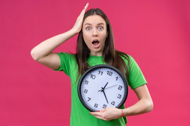 Hermosa joven vestida con camiseta verde sosteniendo el reloj mirando asombrado y sorprendido tocando la cabeza con la mano sobre fondo rosa aislado