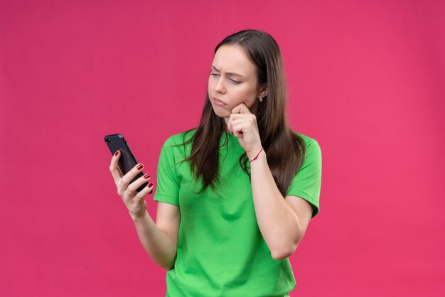 Hermosa joven vestida con camiseta verde sosteniendo smartphone mirando la pantalla con expresión pensativa en la cara de pie sobre fondo rosa aislado