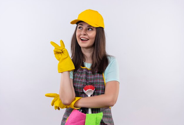 Hermosa joven vestida con delantal, gorra y guantes de goma sonriendo alegremente mirando a un lado mostrando el signo de la victoria