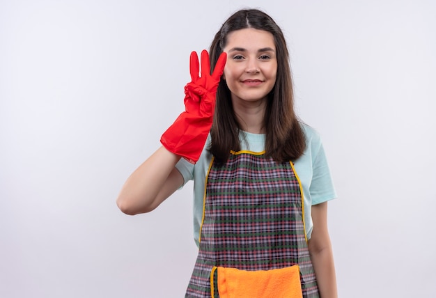 Hermosa joven vestida con delantal y guantes de goma mostrando y apuntando hacia arriba con los dedos número tres sonriendo