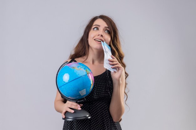 Hermosa joven vestida de lunares con billetes de avión y globo mirando hacia arriba de pie con mirada soñadora sobre fondo blanco.