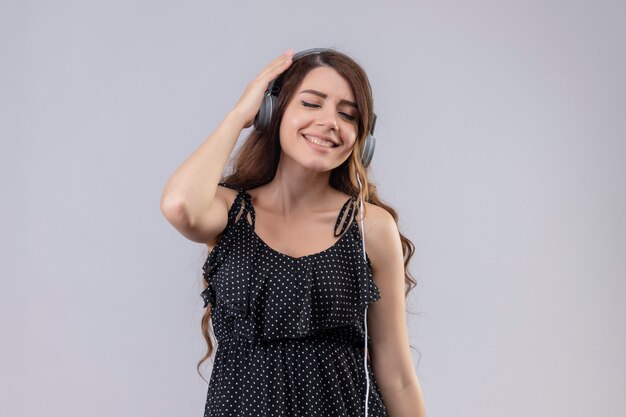 Hermosa joven vestida de lunares disfrutando de su música favorita a través de auriculares inalámbricos de pie con los ojos cerrados sobre fondo blanco.