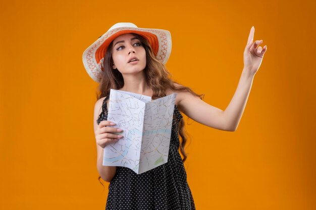 Hermosa joven vestida con lunares en sombrero de verano con mapa gesticulando espere un minuto con seria expresión de confianza en la cara de pie sobre fondo amarillo