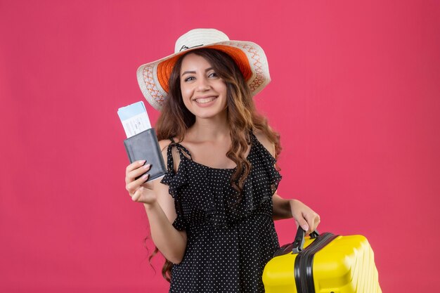 Hermosa joven vestida con lunares en sombrero de verano de pie con maleta sosteniendo boletos de avión mirando a cámara sonriendo alegremente sobre fondo rosa