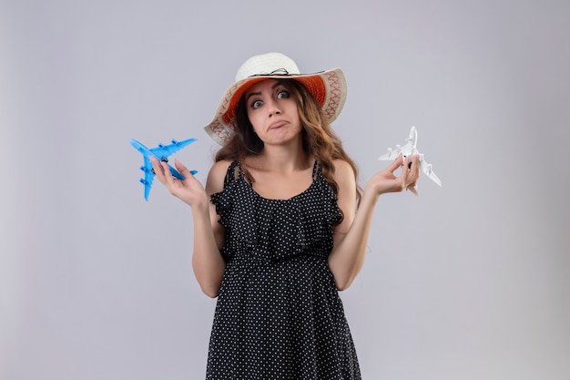 Hermosa joven vestida con lunares en sombrero de verano sosteniendo aviones de juguete despistado y confundido mirando a la cámara de pie con los brazos levantados sin respuesta sobre fondo blanco