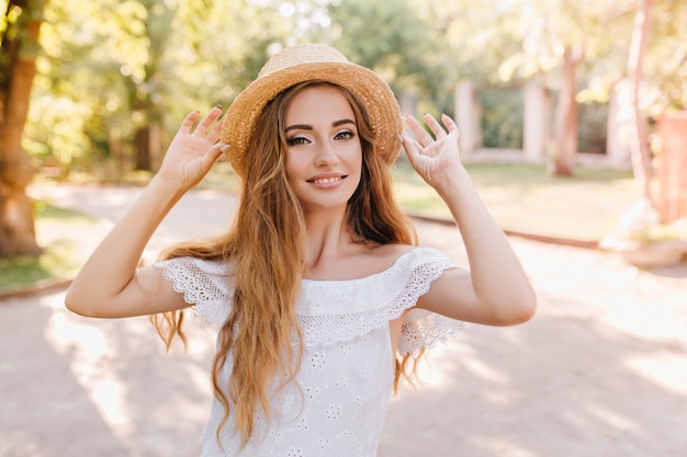 Foto gratuita hermosa joven en vestido blanco de moda caminando en el parque en la mañana soleada y sonriendo. retrato al aire libre de una chica muy de moda posando con placer en sombrero de paja.