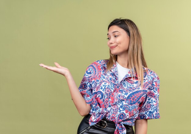 Una hermosa joven vistiendo camisa estampada de paisley levantando la mano y mirándola en una pared verde