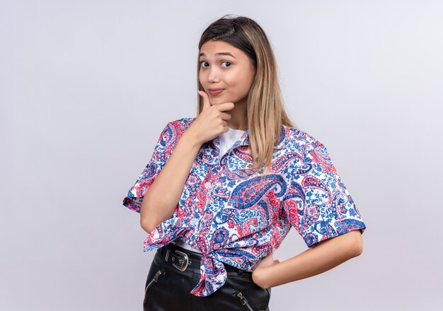 Una hermosa joven vistiendo una camiseta estampada de paisley manteniendo su mano en la barbilla mientras mira en una pared blanca