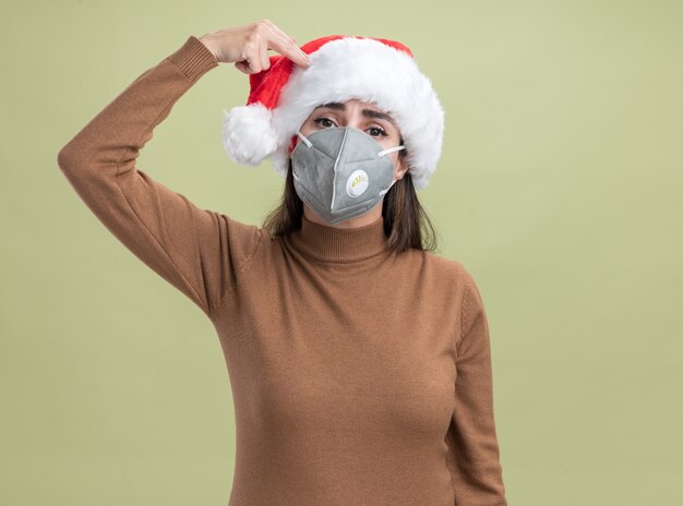 Hermosa joven vistiendo gorro de navidad con máscara médica que muestra el suicidio con gesto de pistola aislado en la pared verde oliva