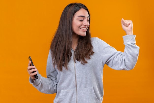 Hermosa jovencita con capucha sosteniendo teléfono mirando a otro lado con placer de pie sobre fondo naranja