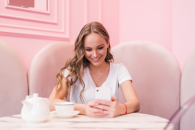 Hermosa jovencita charlando en su teléfono inteligente mientras se relaja en la cafetería después de caminar durante el fin de semana de verano