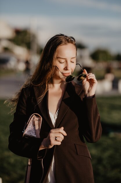 Hermosa jovencita en elegantes gafas de sol y con un bolso de moda al atardecer.