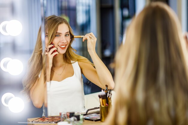 Hermosa jovencita haciendo maquillaje frente al espejo y hablando por teléfono móvil.