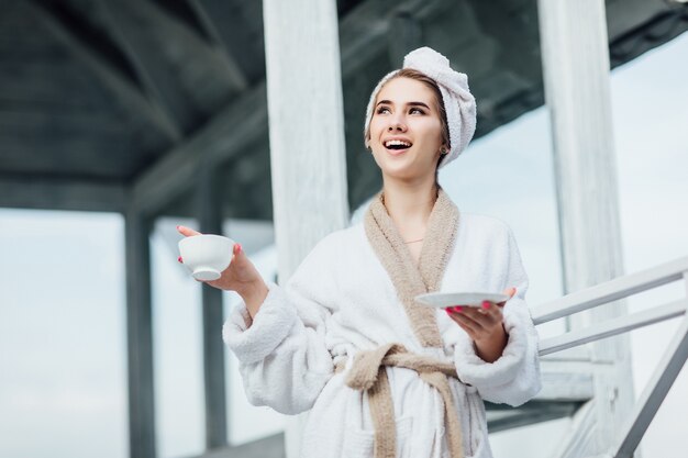 Hermosa y linda joven vestida con túnica blanca permanecer en la terraza de lujo con una taza de café. Vacaciones en las montañas.