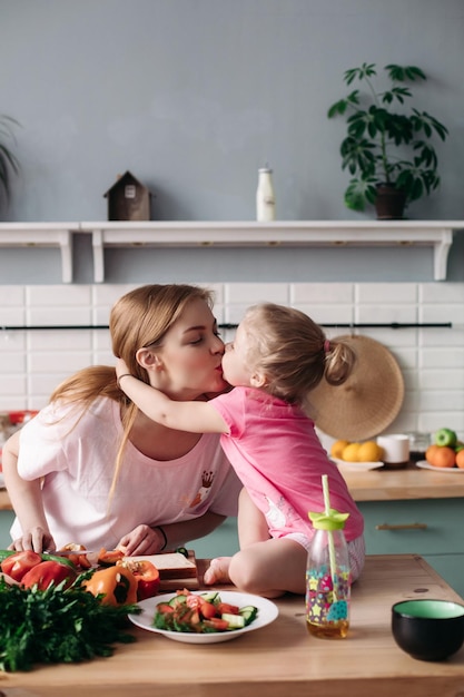 Hermosa madre cocinando la cena en la cocina con su pequeño y lindo hijo Hija sentada en la mesa y comiendo mientras la madre corta pimienta Mujer feliz haciendo el almuerzo de verduras junto con el niño