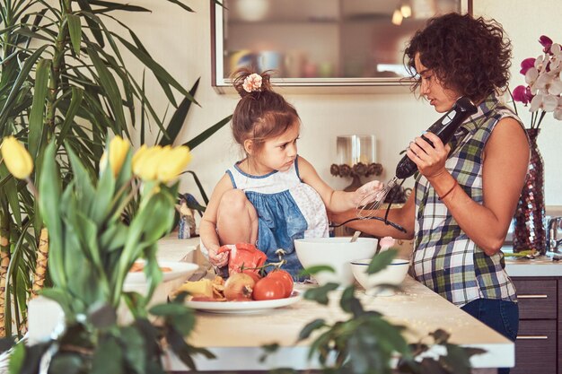 Hermosa madre hispana rizada le enseña a su linda hijita a preparar pizza en la cocina.