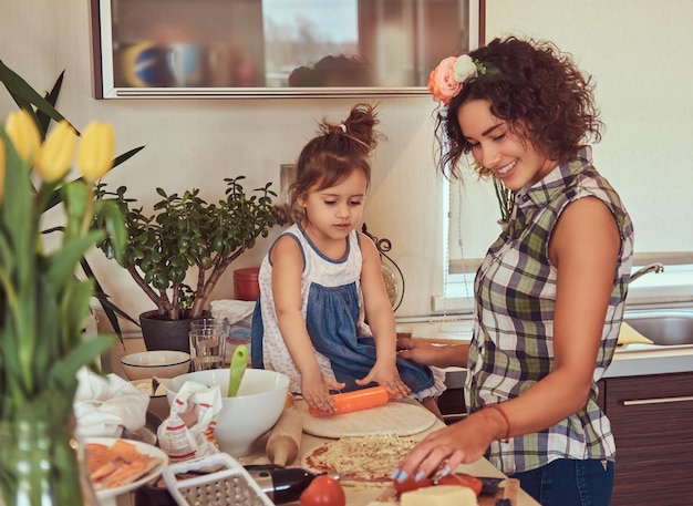 Hermosa madre hispana rizada le enseña a su linda hijita a preparar pizza en la cocina.