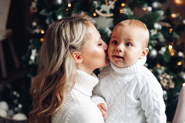 hermosa madre rubia besando la mejilla de su hijo contra el árbol de Navidad.