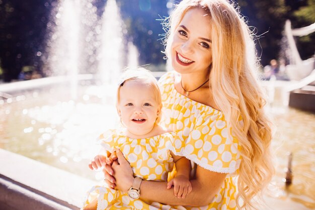 Hermosa madre rubia en vestido manchado amarillo se sienta con su hija en los rayos del sol de la mañana