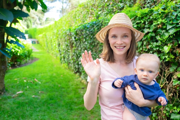 Hermosa madre con sombrero saludando, sosteniendo recién nacido, sonriendo y mirando a cámara. Adorable bebé en manos de mamá mirando seriamente. Tiempo de verano en familia, jardín