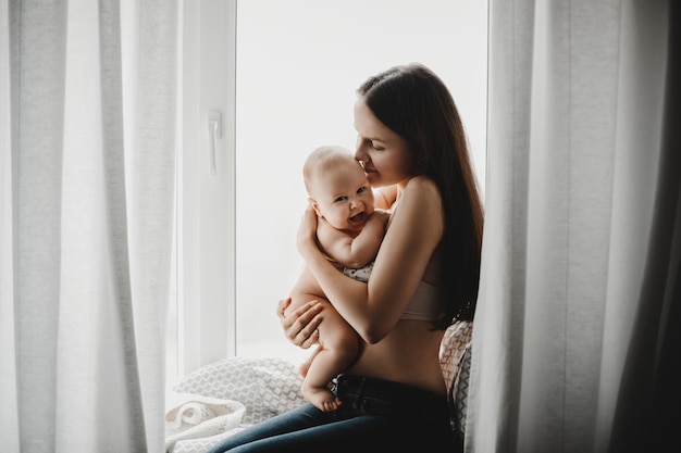 Hermosa madre sostiene feliz niño recién nacido de pie ante una ventana brillante