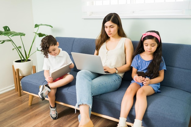 Hermosa mamá escribiendo en la laptop y trabajando mientras se sienta en el sofá con su hijo y su hija. Niños pequeños jugando con una tableta y un teléfono inteligente