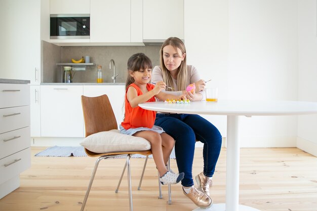 Hermosa mamá pintando huevos de Pascua con hija en la cocina.