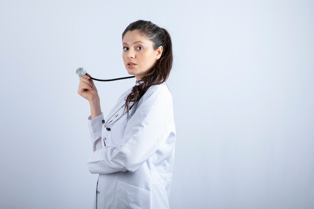 Foto gratuita hermosa médico en bata blanca de pie y posando sobre una pared blanca.