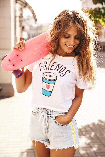 Hermosa modelo de adolescente rubia linda sin maquillaje en ropa blanca hipster de verano con patín rosa centavo posando en el fondo de la calle