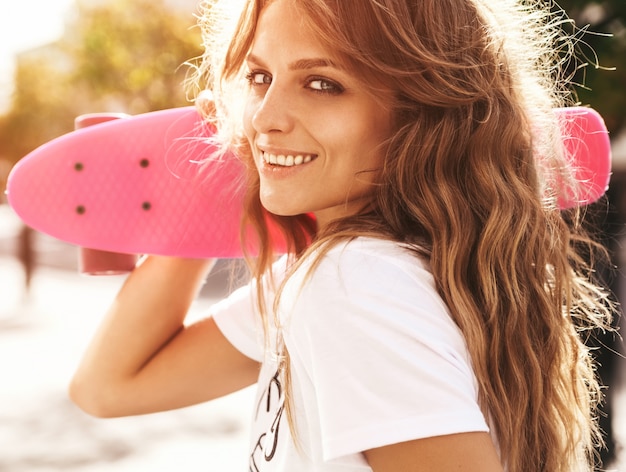 Hermosa modelo de adolescente rubia linda y sonriente sin maquillaje en ropa blanca hipster de verano con patineta rosa centavo posando en el fondo de la calle