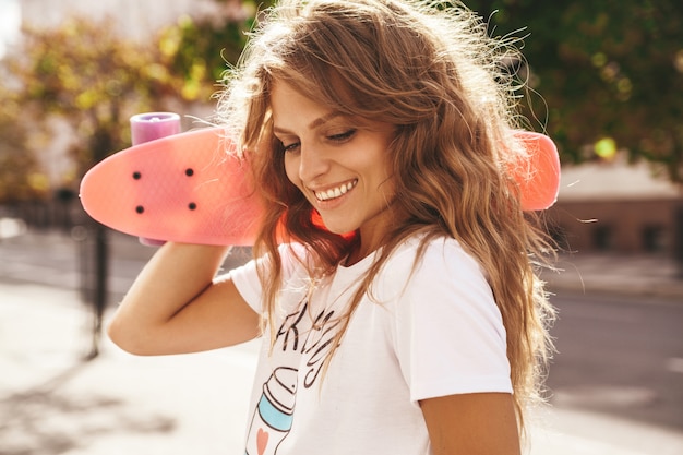 Hermosa modelo de adolescente rubia linda y sonriente sin maquillaje en ropa blanca hipster de verano con patineta rosa centavo posando en el fondo de la calle