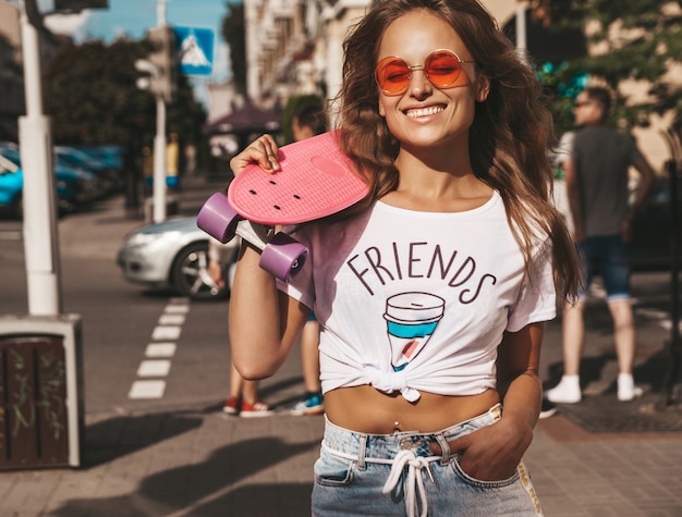 Hermosa modelo de adolescente rubia linda y sonriente sin maquillaje en ropa blanca hipster de verano con patineta rosa centavo posando en el fondo de la calle