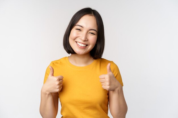 Hermosa modelo asiática sonriente mostrando los pulgares hacia arriba y luciendo complacida recomendando expresar comentarios positivos de pie sobre fondo blanco.