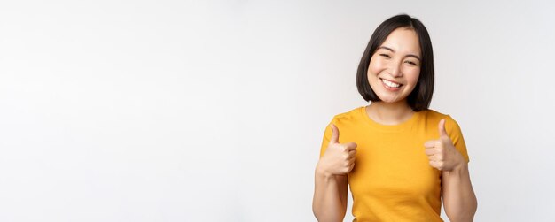 Hermosa modelo asiática sonriente mostrando los pulgares hacia arriba y luciendo complacida recomendando expresar comentarios positivos de pie sobre fondo blanco.