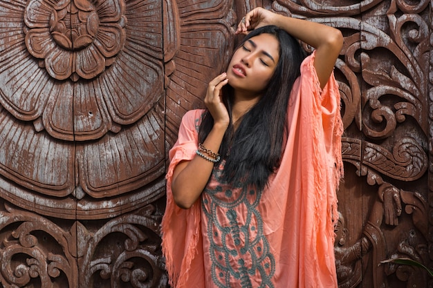 Hermosa modelo asiática en vestido rosa boho posando sobre pared ornamental de madera.