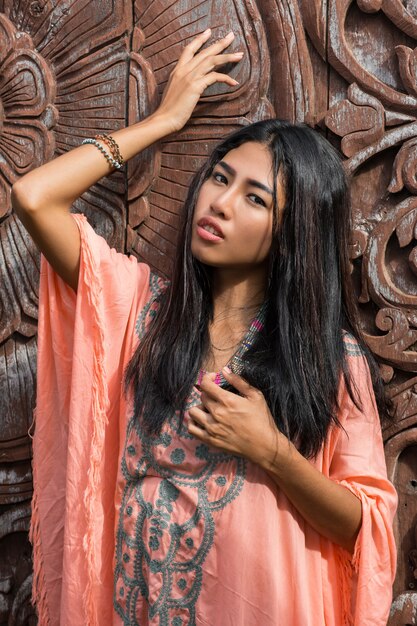 Hermosa modelo asiática en vestido rosa boho posando sobre pared ornamental de madera.