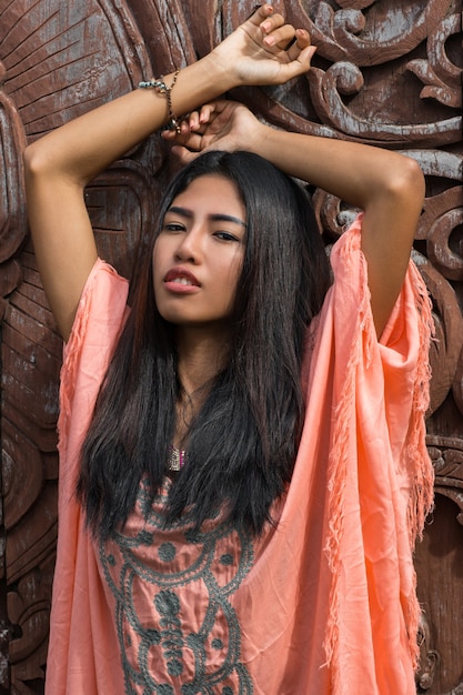 Hermosa modelo asiática en vestido rosa boho posando sobre pared ornamental de madera.