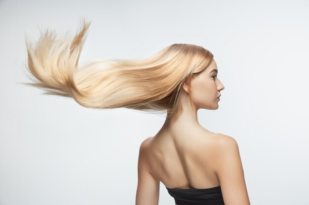 Hermosa modelo con cabello rubio largo, liso y volador aislado sobre fondo blanco de estudio. Modelo caucásico joven con piel bien cuidada y cabello soplando en el aire.