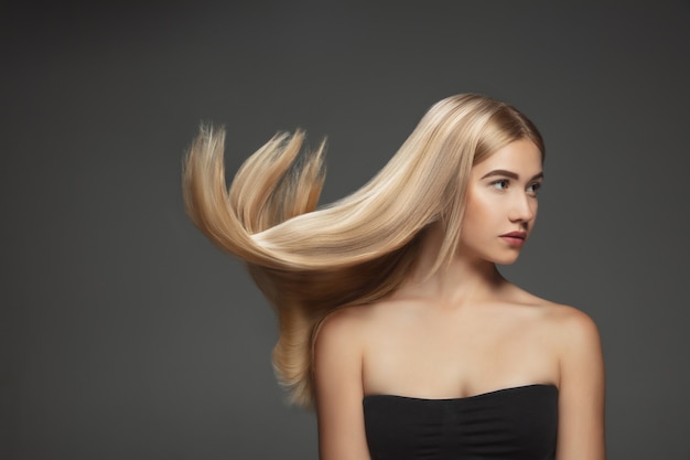 Hermosa modelo con cabello rubio largo, liso y volador aislado sobre fondo gris oscuro de estudio. Modelo caucásico joven con piel bien cuidada y cabello soplando en el aire.