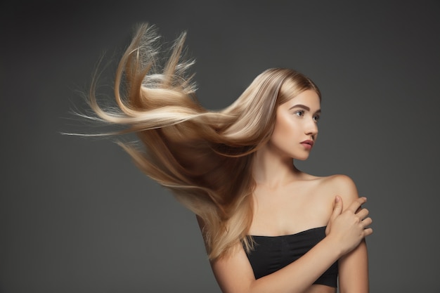 Hermosa modelo con cabello rubio largo, liso y volador aislado sobre fondo gris oscuro de estudio. Modelo caucásico joven con piel bien cuidada y cabello soplando en el aire.