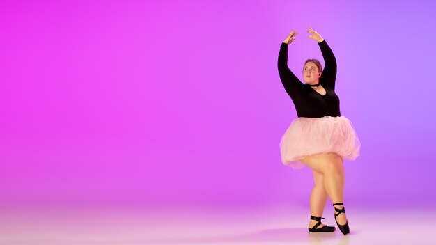 Hermosa modelo caucásica de talla grande practicando ballet sobre fondo degradado de estudio de color rosa púrpura en luz de neón. Concepto de motivación, inclusión, sueños y logros. Vale la pena ser bailarina.