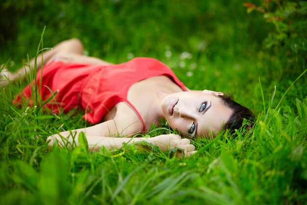 Hermosa modelo de chica morena femenina tumbada en verano verde hierba brillante en el parque con maquillaje en vestido rojo