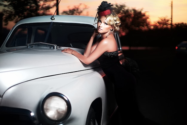 Hermosa modelo de chica rubia de moda sexy con maquillaje brillante y peinado rizado en estilo retro sentado en un auto viejo