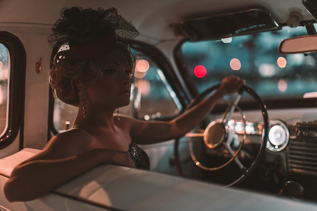 Hermosa modelo de chica rubia de moda sexy con maquillaje brillante y peinado rizado en estilo retro sentado en un auto viejo