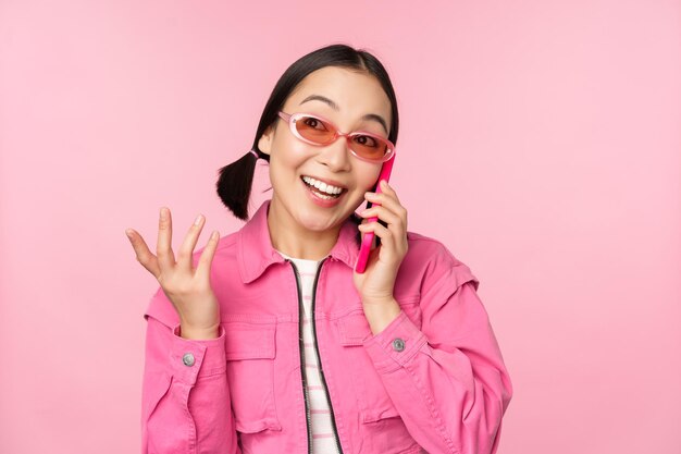 Hermosa modelo coreana con gafas de sol hablando por teléfono móvil con cara feliz usando el servicio celular para llamar a un amigo en el teléfono inteligente de pie sobre fondo rosa