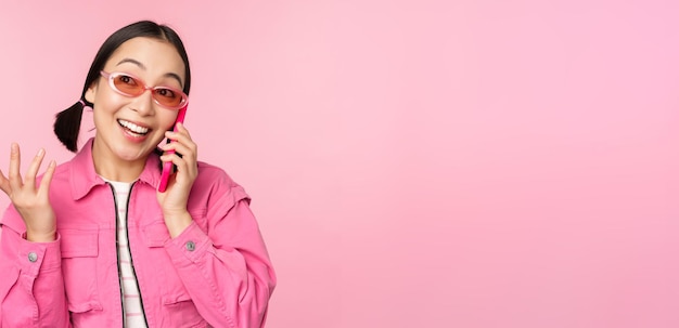 Hermosa modelo coreana con gafas de sol hablando por teléfono móvil con cara feliz usando el servicio celular para llamar a un amigo en el teléfono inteligente de pie sobre fondo rosa