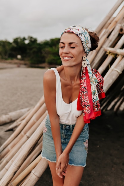 Hermosa modelo en falda de mezclilla y chal en la cabeza posando en la playa cerca del océano con una sonrisa maravillosa