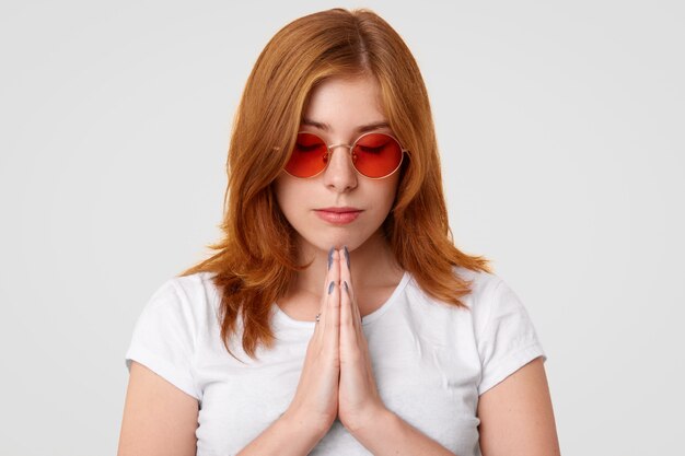 Hermosa modelo femenina centrada mantiene las manos en gesto de oración, le pide a Dios el bienestar de su familia, usa gafas de sol modernas