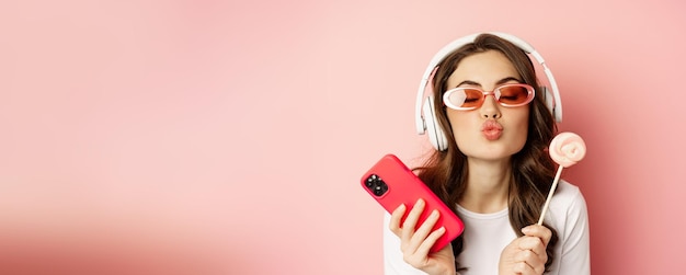 Foto gratuita hermosa modelo femenina escuchando música en auriculares con piruleta y teléfono móvil posando en cantado